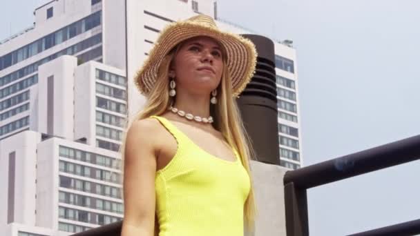穿着黄色泳衣 头戴太阳帽的年轻女子在城市屋顶无限大池边行走的中等慢镜头跟踪 — 图库视频影像