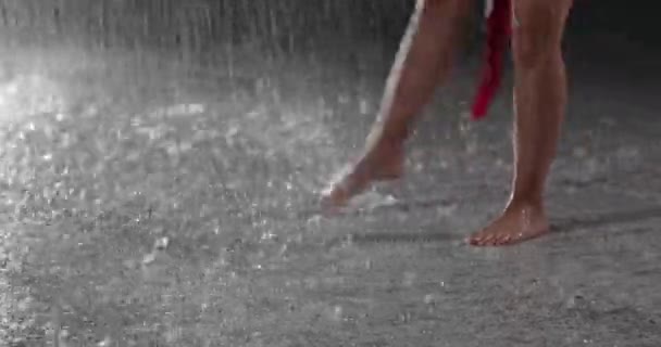 赤いドレスで若い女性ダンサーを誘惑の足のビュースポットライトに対して暗いスタジオで雨の中で裸足で実行 床に落ちる水滴雨のシャワーを模倣 — ストック動画
