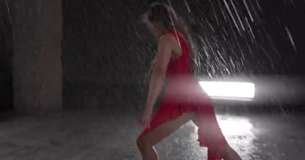 赤いドレスで若い女性ダンサーを誘惑上のスポットライトに対してスタジオで雨の中で実行 床に落ちる水滴雨のシャワーを模倣します カメラはダンサーに続く カメラレンズの水滴 — ストック動画