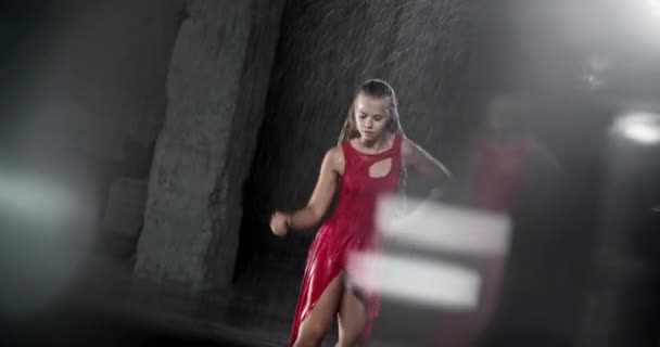 赤いドレスで若い女性ダンサーを誘惑上のスポットライトに対して暗いスタジオで雨の中で実行 床に落ちる水滴雨のシャワーを模倣します 光の反射によるサイドビュー — ストック動画