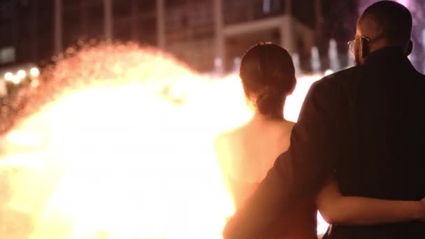 ライトアップされた噴水と夜のモダンなガラスの建物の前で抱き合う愛情のあるカップルの背景 スローモーション 中棒アーチショット — ストック動画