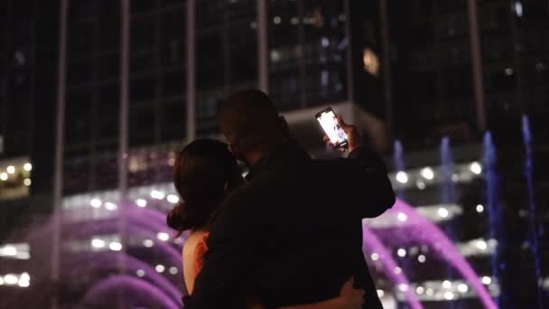 夜のカラフルな噴水ショーの前で抱き合う愛情のあるカップルの背景 彼らはスマートフォーンでセルフィーを作る スローモーションボケドリーアウトショット — ストック動画