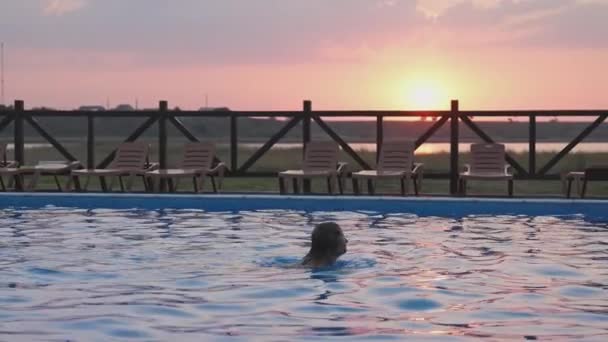 Chica con el pelo mojado en bikini de color emerge de la piscina con agua transparente y transparente sobre el fondo de la puesta de sol de verano — Vídeos de Stock