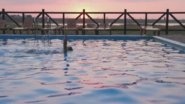 Woman swims in a pool with clear water on the background of a summer sunset on vacation — Stock Video