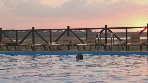 Frau schwimmt in einem Pool mit klarem Wasser vor dem Hintergrund eines sommerlichen Sonnenuntergangs im Urlaub — Stockvideo