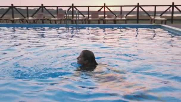 Woman swims in a pool with clear water on the background of a summer sunset on vacation — Stock Video