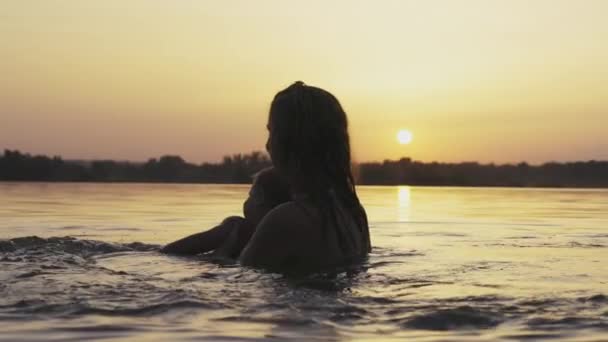 Anya játszik egy meztelen baba oversleeves a tóban a háttérben a nyári naplemente — Stock videók