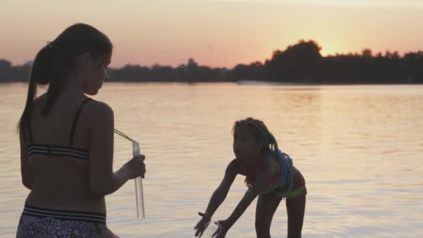 Забавная девушка ловит пузырьки на берегу озера, которые ее старшая сестра отпускает — стоковое видео