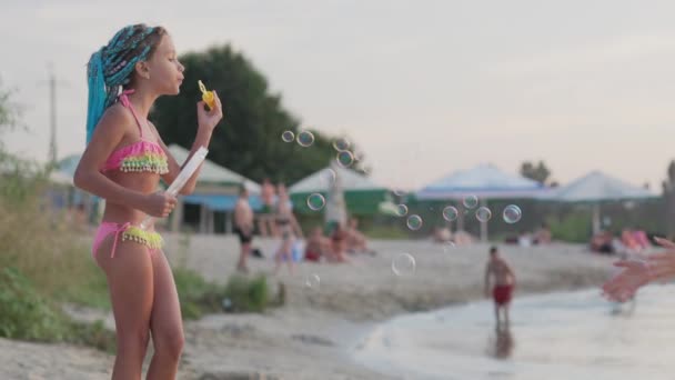 Молодая девушка ловит пузырьки на берегу озера, которые ее младшая сестра отпускает — стоковое видео