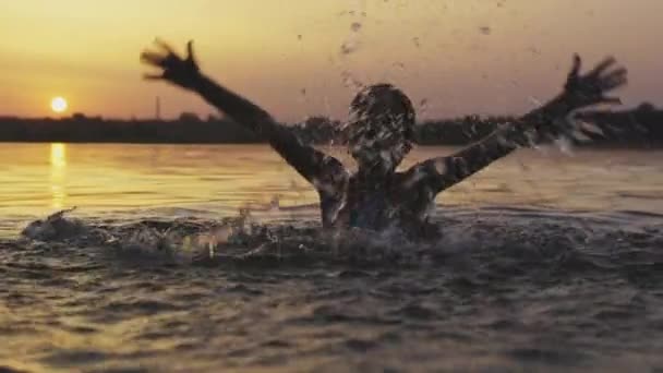 A girl plays with water in the lake and splashes it to the sides against the backdrop of a sunset — Stock Video