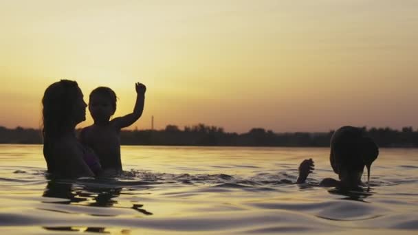 Mãe com o bebê que ela segura em seus braços brincando com sua filha e espirrando no lago contra o fundo do pôr do sol — Vídeo de Stock