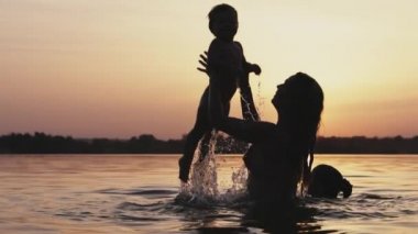 Annem gölde kolsuz çıplak bir bebekle oynuyor yaz günbatımının arka planında.
