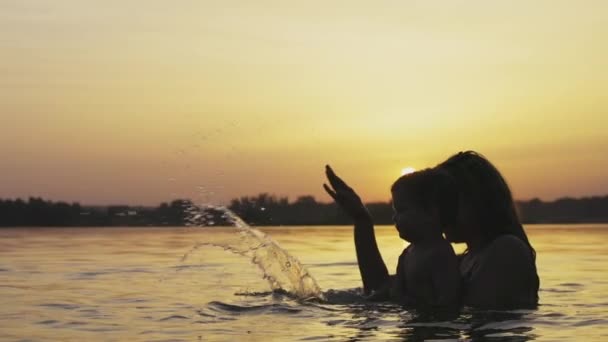 Moeder met de baby ze houdt in haar armen spelen met haar dochter en spetteren in het meer tegen de achtergrond van zonsondergang — Stockvideo