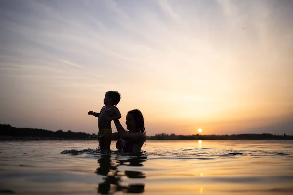 Mom plays with a naked baby in oversleeves in the lake against the background of a summer sunset — Stock Photo, Image