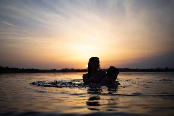 Η μαμά παίζει με ένα γυμνό μωρό με μανίκια στη λίμνη στο φόντο ενός καλοκαιρινού ηλιοβασιλέματος. — Φωτογραφία Αρχείου