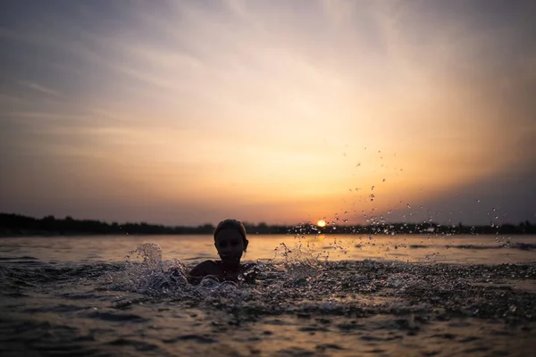 En flicka leker med vatten i sjön och stänk den åt sidorna mot bakgrund av en solnedgång — Stockfoto