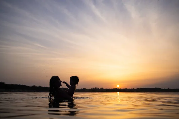 Mamma leker med en naken baby i oversleeves i sjön mot bakgrund av en sommar solnedgång — Stockfoto