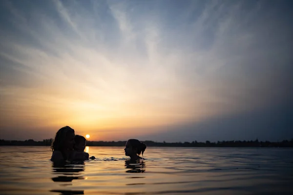 Mamma med barnet hon håller i sina armar leker med sin dotter och stänk i sjön mot bakgrund av solnedgången — Stockfoto
