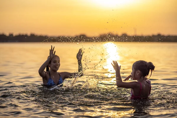 Deux sœurs éclaboussant l'eau jouant dans le lac au coucher du soleil fond — Photo