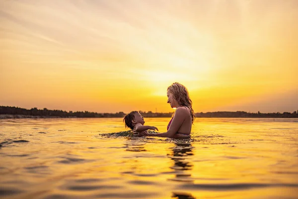 Mom plays with a naked baby in oversleeves in the lake against the background of a summer sunset — Stock Photo, Image