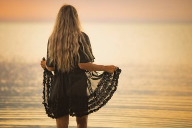 Siyah bikinili, sarı şık saçlı ve dantel şallı bir kız nehir boyunca yürüyor.