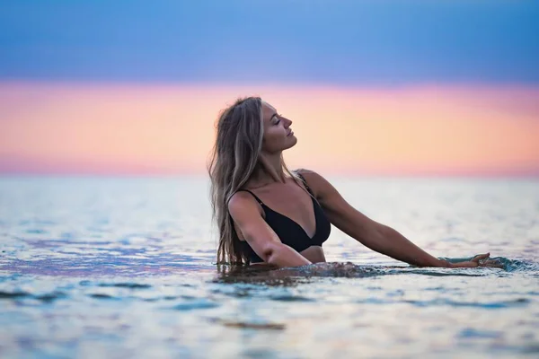 Une fille aux cheveux blonds en maillot de bain noir éclabousse les côtés alors qu'elle est assise dans un estuaire sur un fond de coucher de soleil — Photo