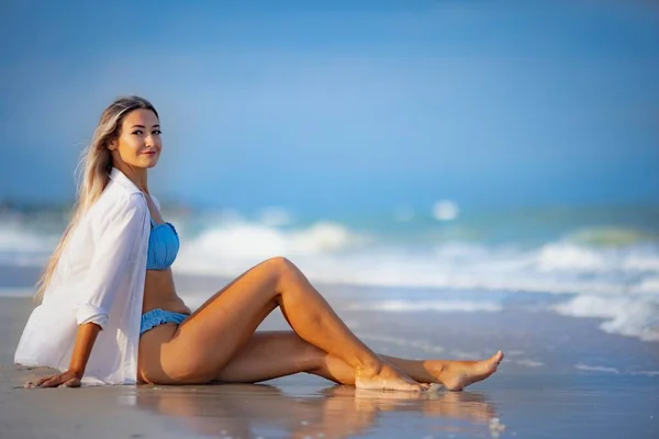 Una chica en un traje de baño azulado y camisa se sienta cerca del borde del agua y mira hacia el horizonte —  Fotos de Stock