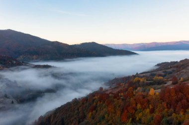 Terracotta ve yeşil ağaçlarla kaplı dağlar, parlak mavi bulutsuz gökyüzü altında büyüyorlar. Beyaz sisli panorama manzarasının kalın tabakasından dışarı çıkıyorlar.
