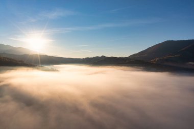 Güneş, parlak mavi gökyüzünde gün batımında kalın kabarık bulutların üzerinde parlıyor. Arkaplanda dağlar var.