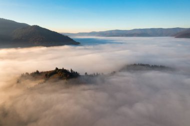 Terracotta ve yeşil ağaçlarla kaplı dağlar, parlak mavi bulutsuz gökyüzü altında büyüyorlar. Beyaz sisli panorama manzarasının kalın tabakasından dışarı çıkıyorlar.