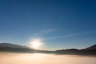 Güneş, parlak mavi gökyüzünde gün batımında kalın kabarık bulutların üzerinde parlıyor. Arkaplanda dağlar var.