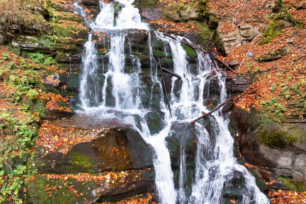 黄色の木々やテラコッタの葉が秋の日に落ちる森の中の山の急な斜面の岩や石に落ちる狭い滝の流れ — ストック写真