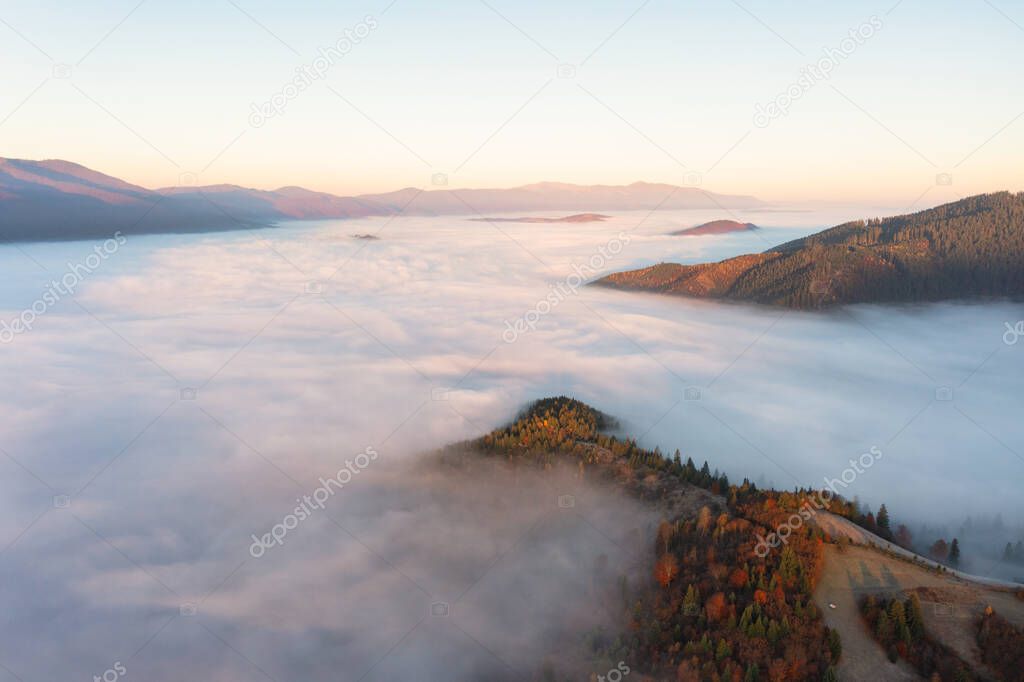 Thick layer of fog covering high mountains with terracotta and green trees growing under bright blue cloudless sky panorama view