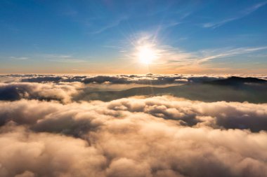 Güneş, parlak mavi gökyüzünde gün batımında kalın kabarık bulutların üzerinde parlıyor. Arkaplanda dağlar var.