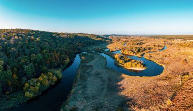 Gölgede yoğun yeşil orman ve günbatımında bulutlu gökyüzünün altında uzun kıvrımlı nehir dalları yakınında geniş çimenli çayır manzarası