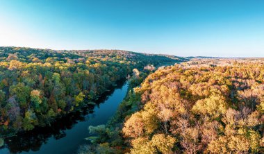 Gölgede yoğun yeşil orman ve günbatımında bulutlu gökyüzünün altında uzun kıvrımlı nehir dalları yakınında geniş çimenli çayır manzarası