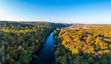 Gölgede yoğun yeşil orman ve günbatımında bulutlu gökyüzünün altında uzun kıvrımlı nehir dalları yakınında geniş çimenli çayır manzarası