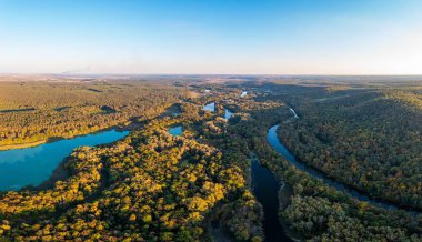 Ormanla kaplı tepeler çimenlerin etrafını sarar ve gün batımında tüylü turuncu bulutların altında koyu mavi sularla dolu uzun kıvrımlı bir nehir.