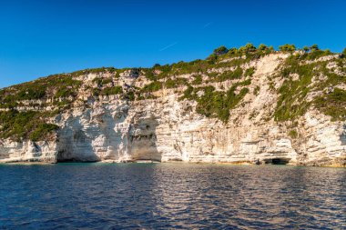 Light-colored layered rock creating cave on beach of Corfu island in Greece with clear turquoise water under blue cloudless sky