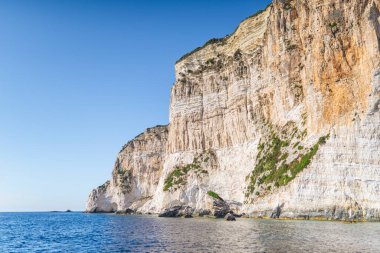 Green forest-covered high rocky steep cliffs on shore of Grecian Corfu island with clear blue sea water under cloudless sky panorama view