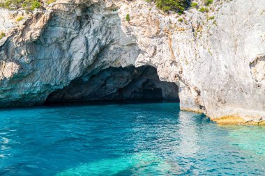 Layered light-colored rocks creating dark cave in sea beach of Corfu island in Greece with clear turquoise water reflecting stones