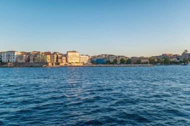 Multistory buildings of historical town on Corfu island shore behind turquoise water under blue cloudy sky lit by pink sunset panorama view
