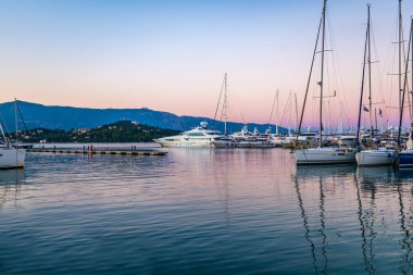 Pier of Grecian Corfu island with numerous white-colored motorboats and yachts moored on water reflecting masts and sky at pink sunset