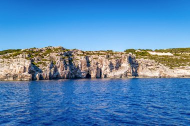 Plant-covered high rocky cliffs on shore of Corfu island in Greece with clear turquoise sea water under blue cloudless sky panorama view