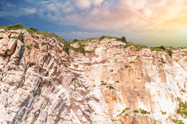 High layered light-colored stoned rocky cliffs covered with plants on Corfu island sea beach with clear blue water under cloudy sky at sunset