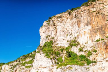 High layered light-colored stoned rocky cliffs covered with plants on Corfu island sea beach with clear blue water under cloudy sky at sunset