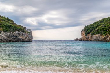 Calm beach and clear turquoise-colored water of Mediterranean sea of Grecian Corfu island near high steep hill covered with forest