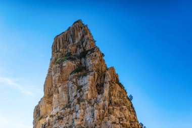 High rocky cliff with plants against cloudy sky at sunset