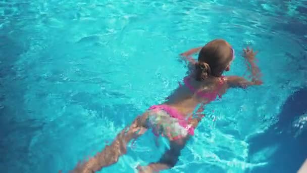 Dziewczyna w różowym stroju kąpielowym w fioletowych okularach unosi się na basenie z błękitną wodą w letni dzień — Wideo stockowe