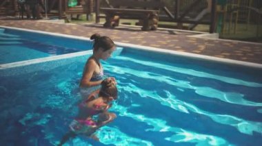 Elder sister in leopard swimsuit pulls little sister out of the pool with blue water